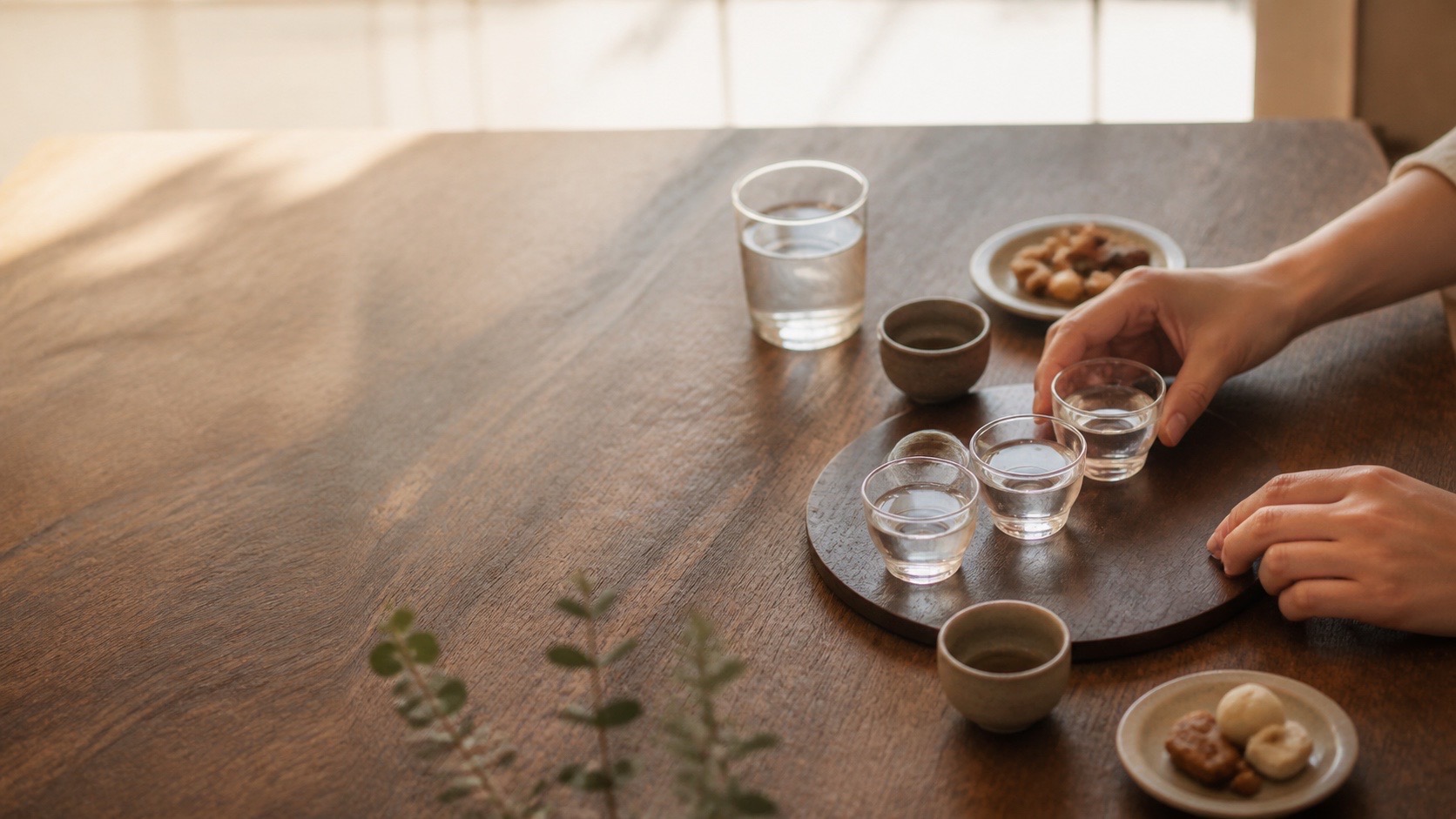 Anonymous hands arranging small unbranded sake-tasting cups beside water and simple snacks